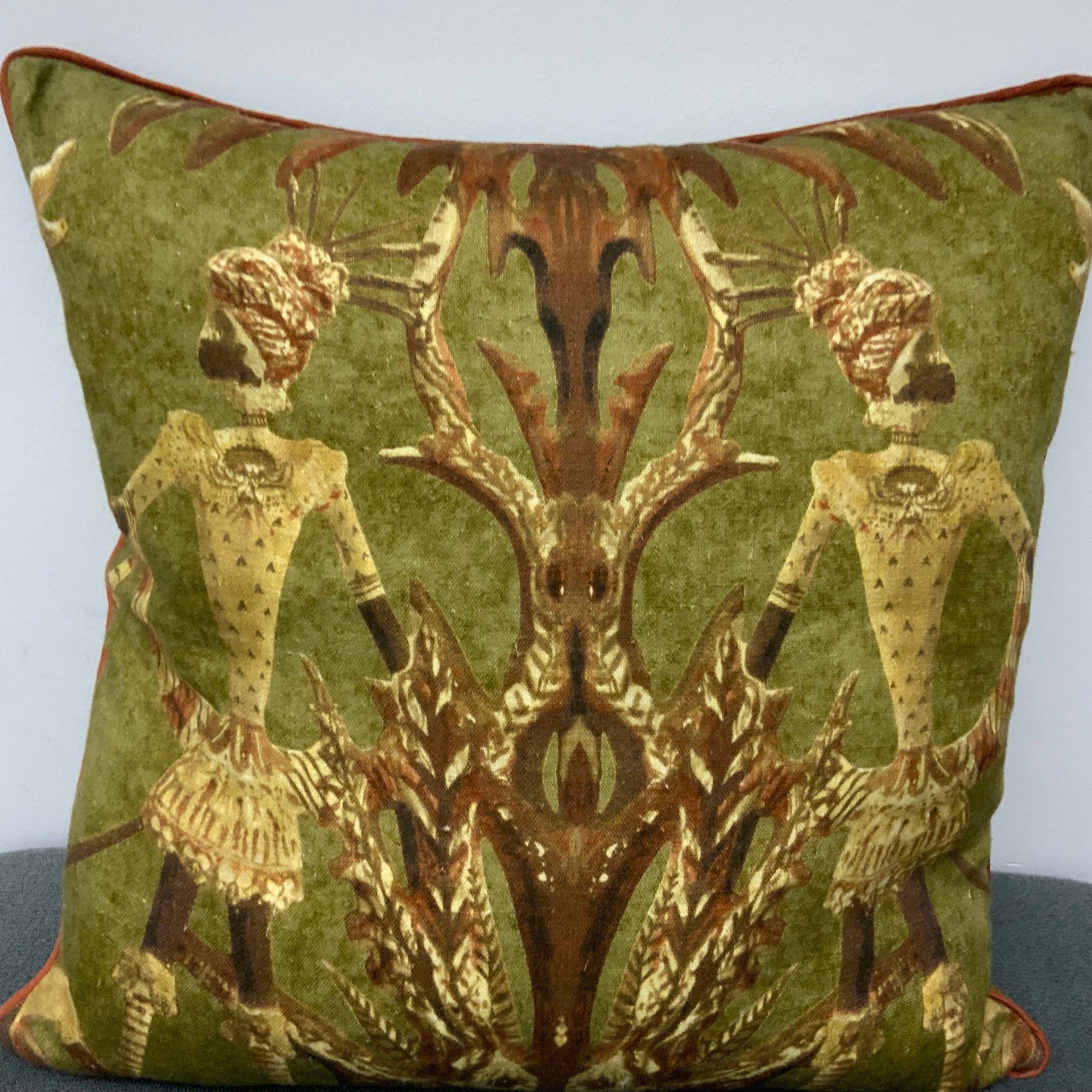 An olive green square decorative pillow featuring a symmetrical, mirror-image design of two stylized tribal figures in traditional headdress. The figures are rendered in ochre and brown tones, framed by abstract botanical patterns. The pillow is finished with a thin terracotta-colored piped edge.