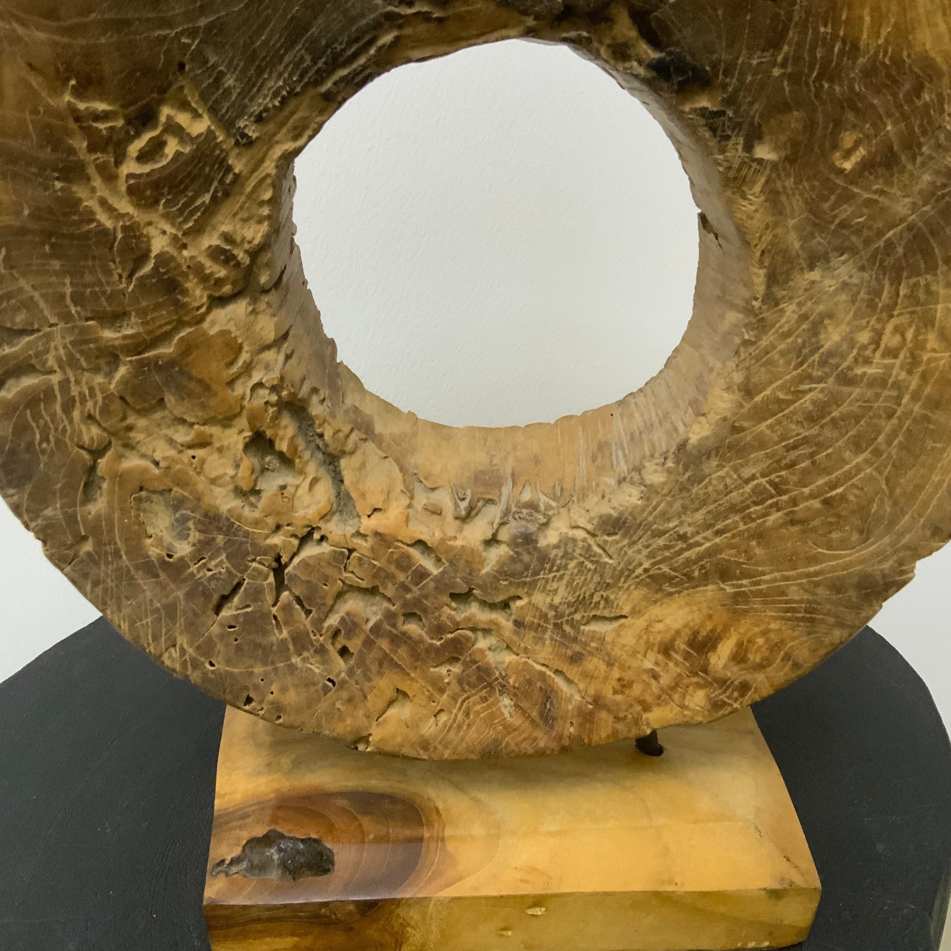 Hand-carved circular wood sculpture with natural grain and weathered textures, mounted on a solid wood base with thin metal rods.