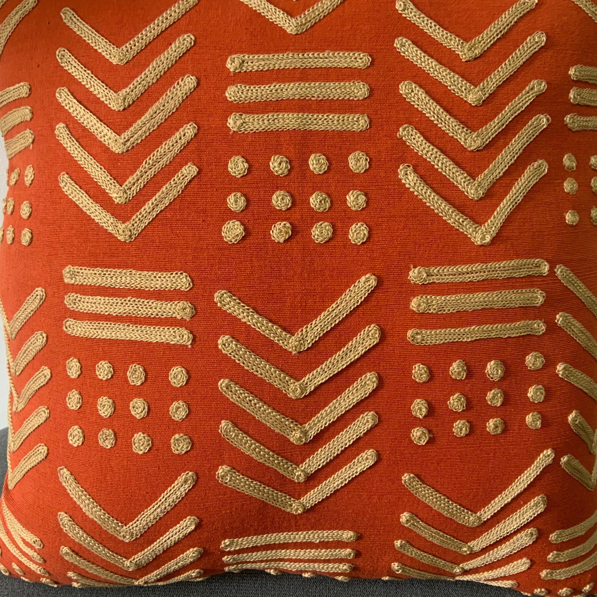 Square terracotta orange throw pillow  with raised gold-toned geometric embroidery in a tribal mudcloth design featuring chevrons and dots.