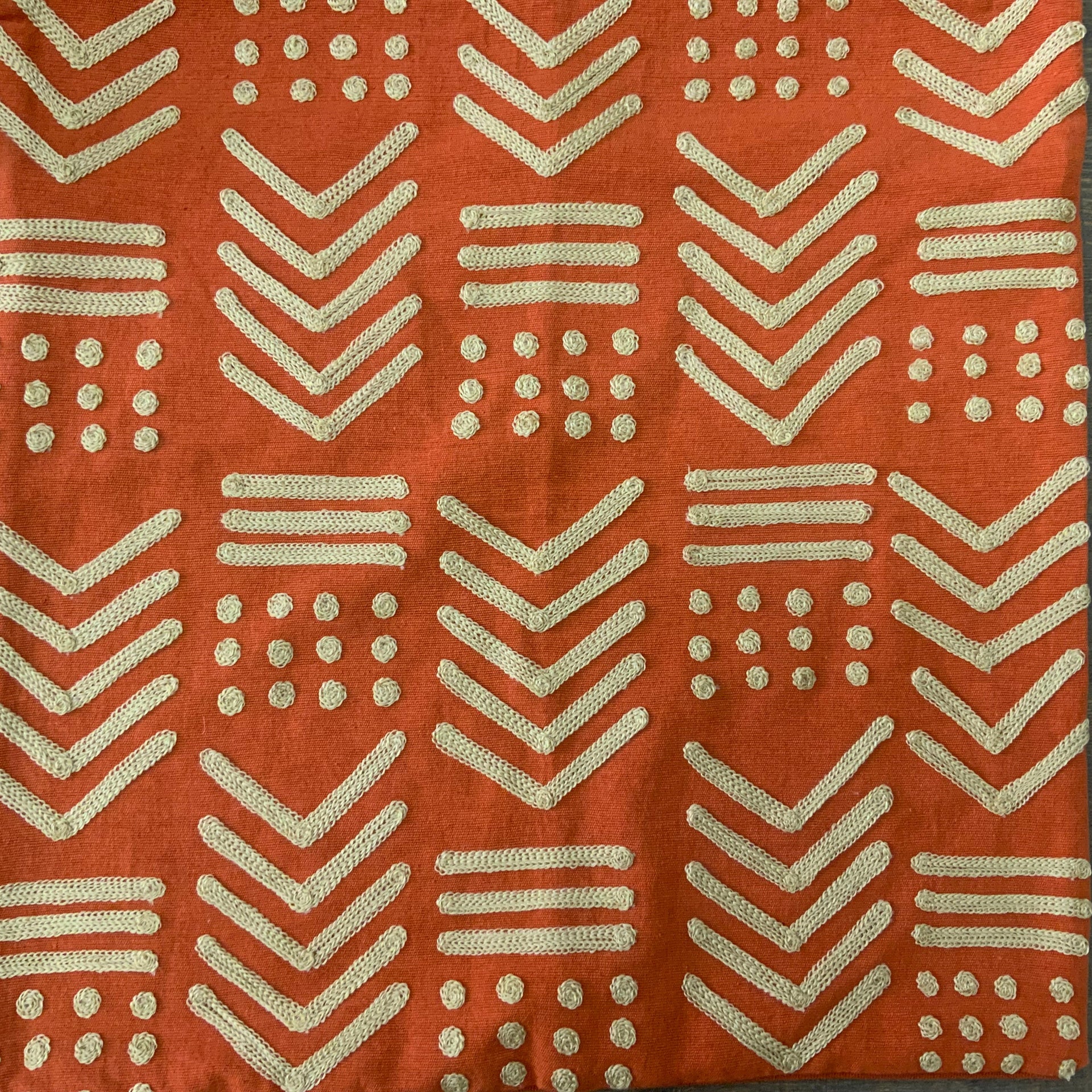 Square terracotta orange throw pillow cover with raised gold-toned geometric embroidery in a tribal mudcloth design featuring chevrons and dots.