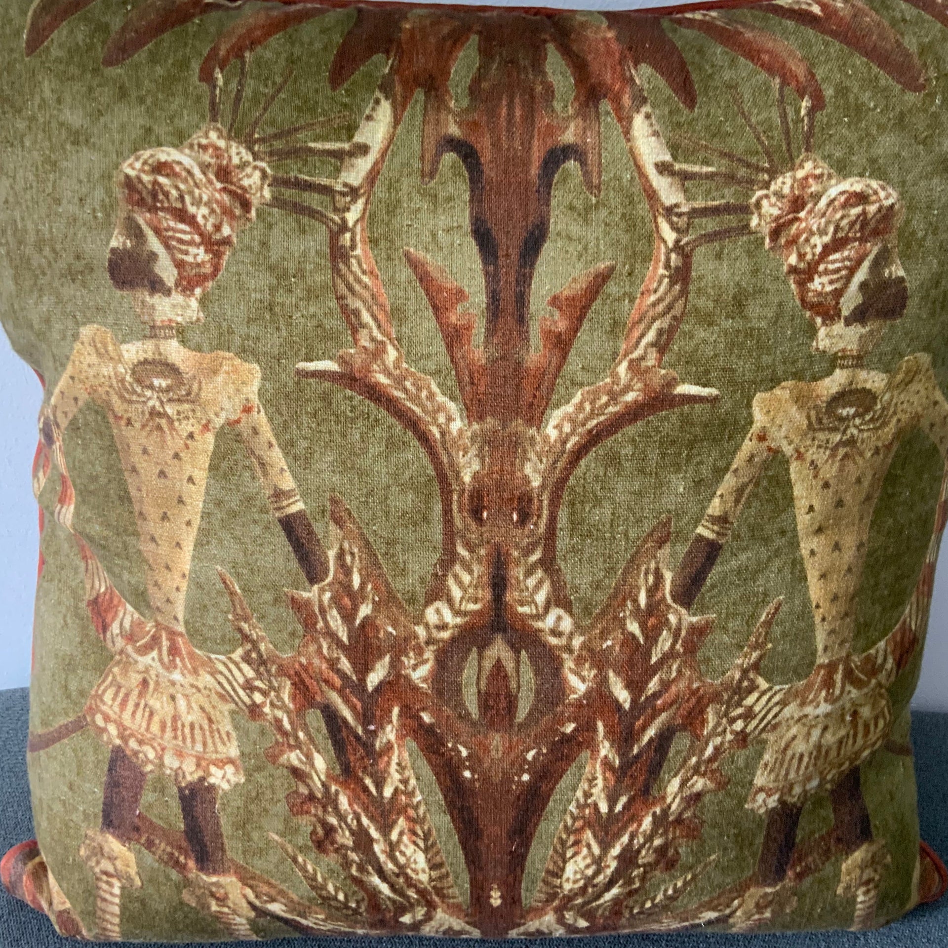 An olive green square decorative pillow featuring a symmetrical, mirror-image design of two stylized tribal figures in traditional headdress. The figures are rendered in ochre and brown tones, framed by abstract botanical patterns. The pillow is finished with a thin terracotta-colored piped edge.