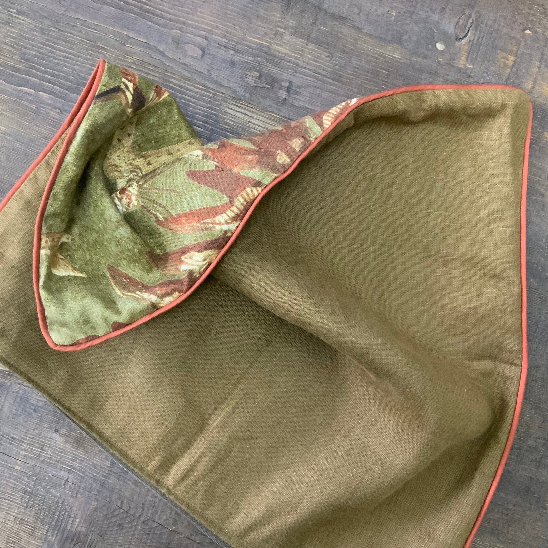 An olive green square decorative pillow featuring a symmetrical, mirror-image design of two stylized tribal figures in traditional headdress. The figures are rendered in ochre and brown tones, framed by abstract botanical patterns. The pillow is finished with a thin terracotta-colored piped edge.