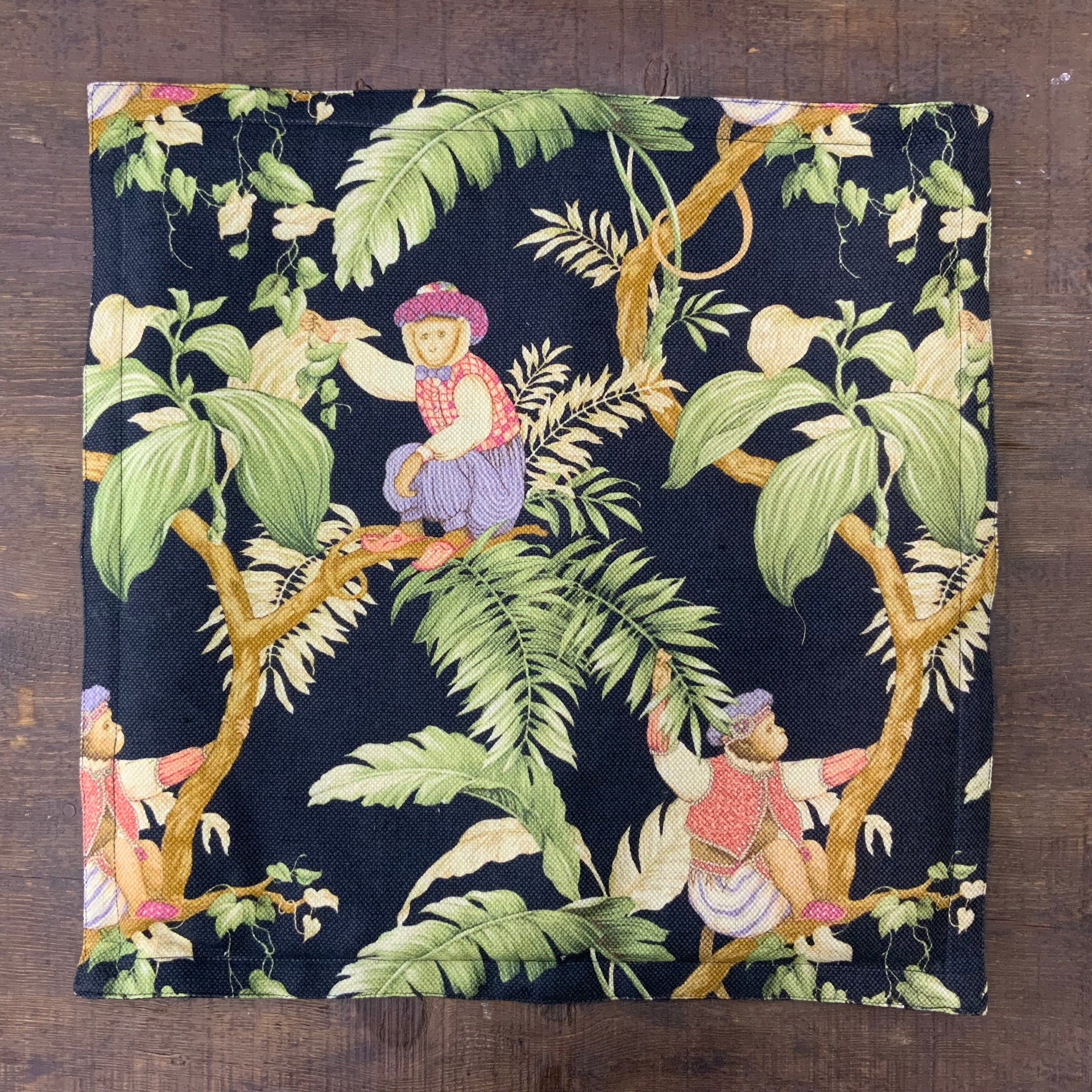 Decorative black throw cushion featuring a tropical jungle print with illustrated monkeys sitting on tree branches surrounded by lush green fern leaves and botanical foliage.
