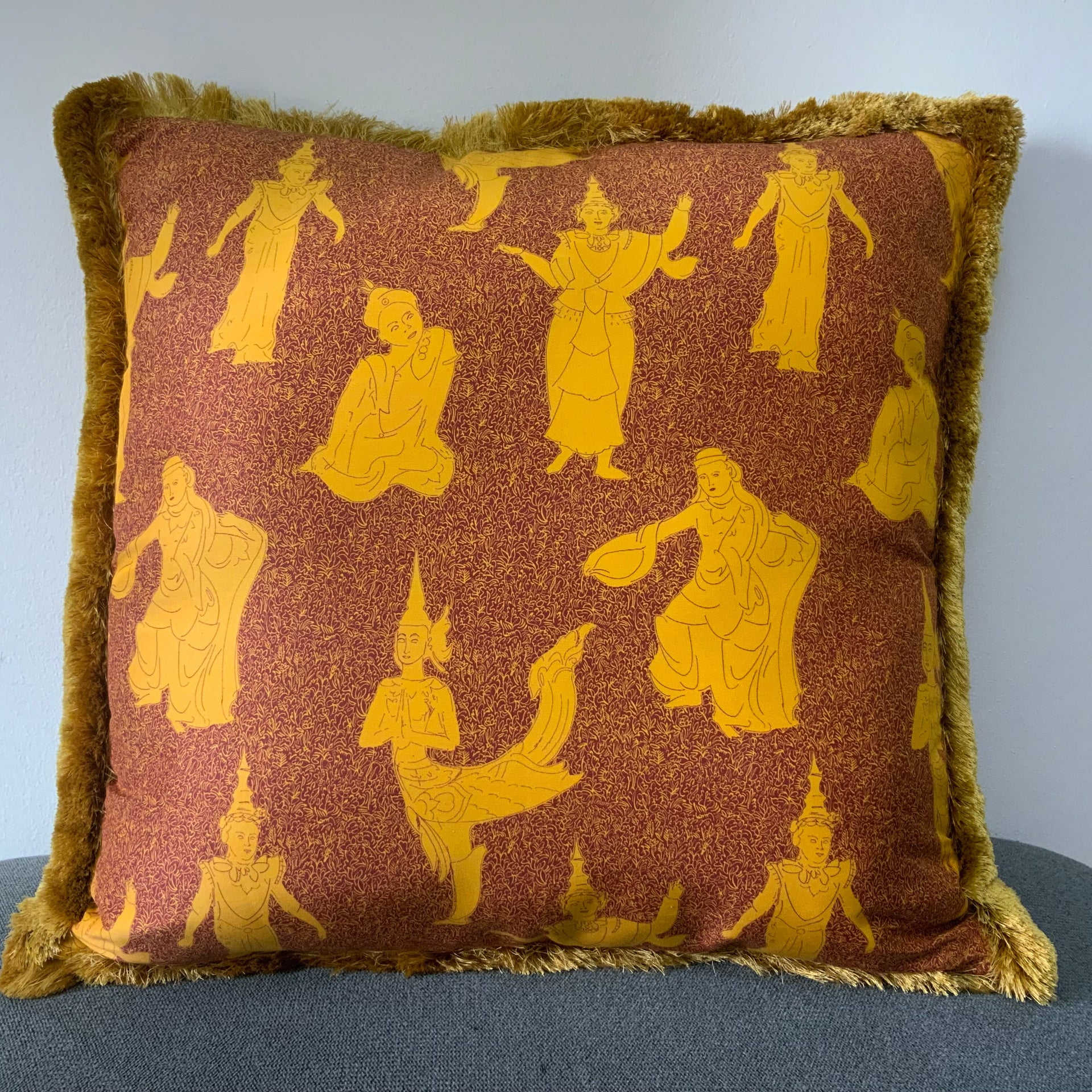 Decorative square pillow cover featuring a traditional Thai celestial dancer pattern in saffron gold on a burnt orange background, finished with a thick golden bullion fringe.