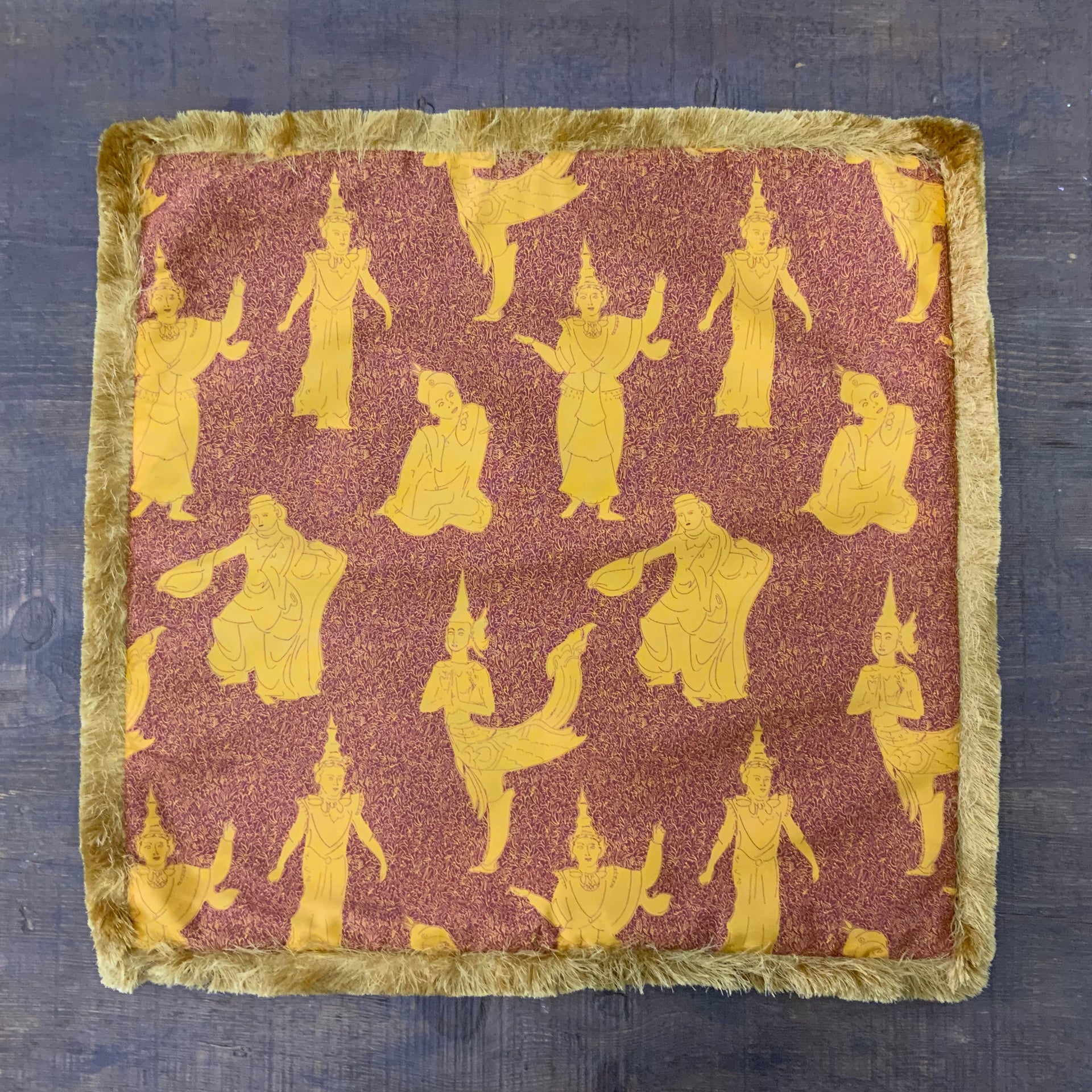 Decorative square pillow cover featuring a traditional Thai celestial dancer pattern in saffron gold on a burnt orange background, finished with a thick golden bullion fringe.