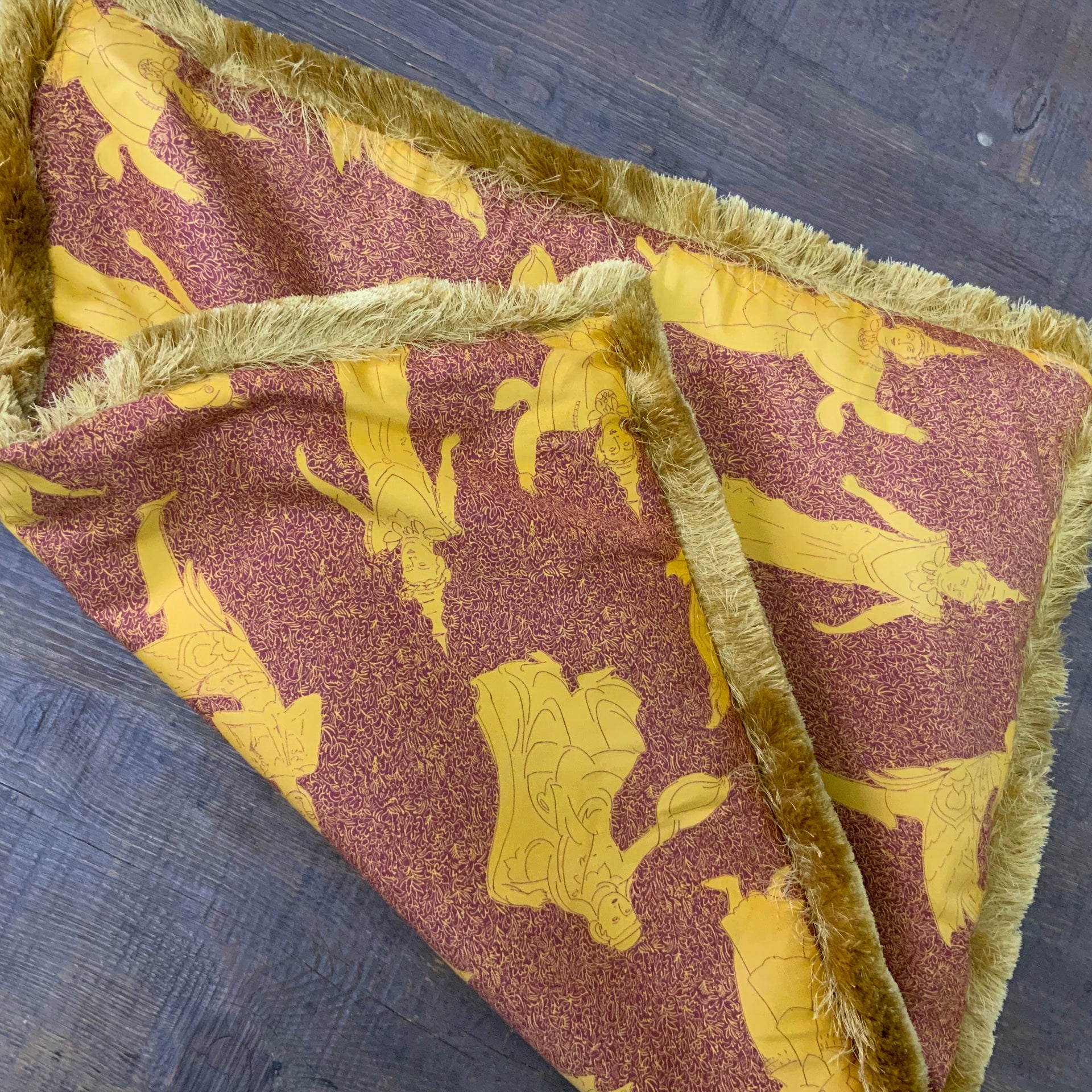 Decorative square pillow cover featuring a traditional Thai celestial dancer pattern in saffron gold on a burnt orange background, finished with a thick golden bullion fringe. Decorative square pillow cover featuring a traditional Thai celestial dancer pattern in saffron gold on a burnt orange background, finished with a thick golden bullion fringe.