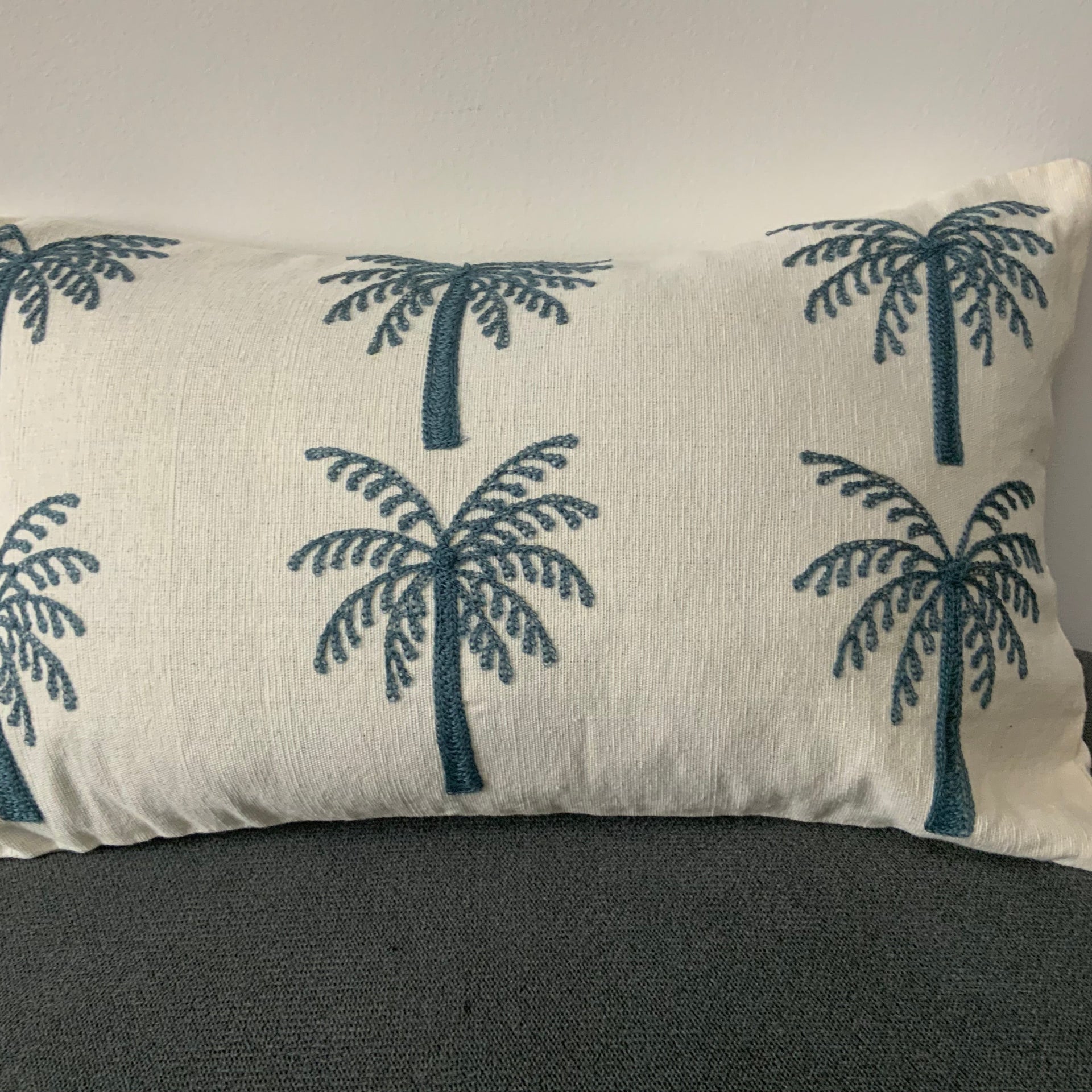 A rectangular off-white lumbar cushion featuring two rows of three identical palm trees embroidered in deep teal blue thread.