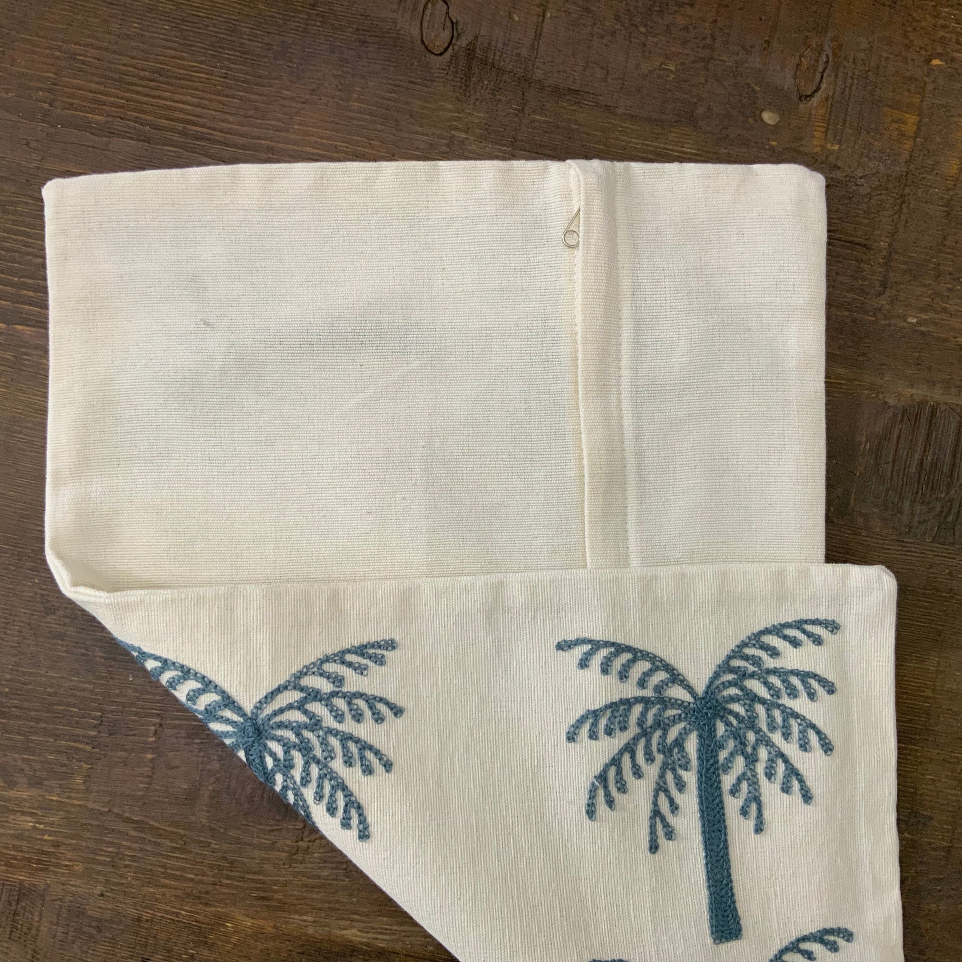A rectangular off-white lumbar cushion featuring two rows of three identical palm trees embroidered in deep teal blue thread.