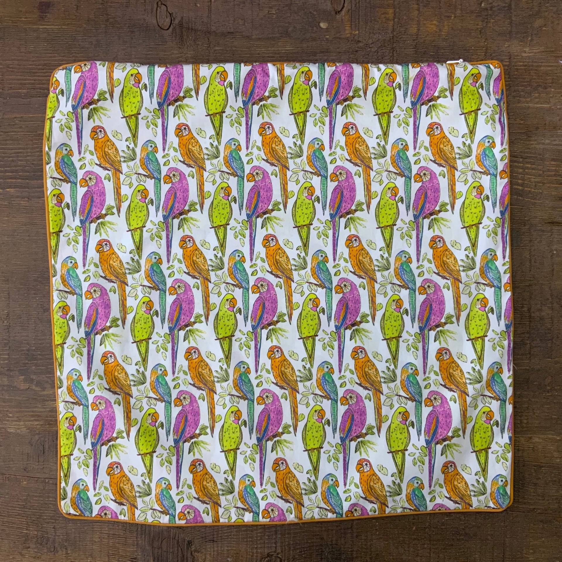 An upright, square decorative pillow cover featuring a vibrant, hand-drawn repeating pattern of tropical parrots perched on thin branches with small green leaves. The parrots are illustrated in a variety of color combinations, including magenta and purple, bright lime green, and amber orange with teal accents. The pillow has a distinct mustard-yellow piped edge.