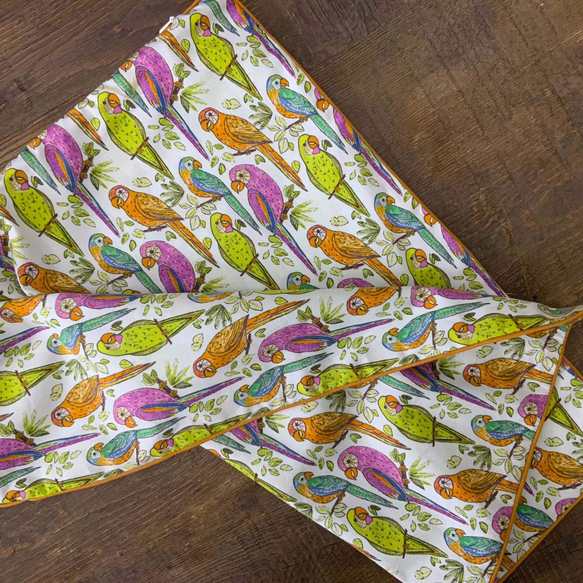 An upright, square decorative pillow cover featuring a vibrant, hand-drawn repeating pattern of tropical parrots perched on thin branches with small green leaves. The parrots are illustrated in a variety of color combinations, including magenta and purple, bright lime green, and amber orange with teal accents. The pillow has a distinct mustard-yellow piped edge.