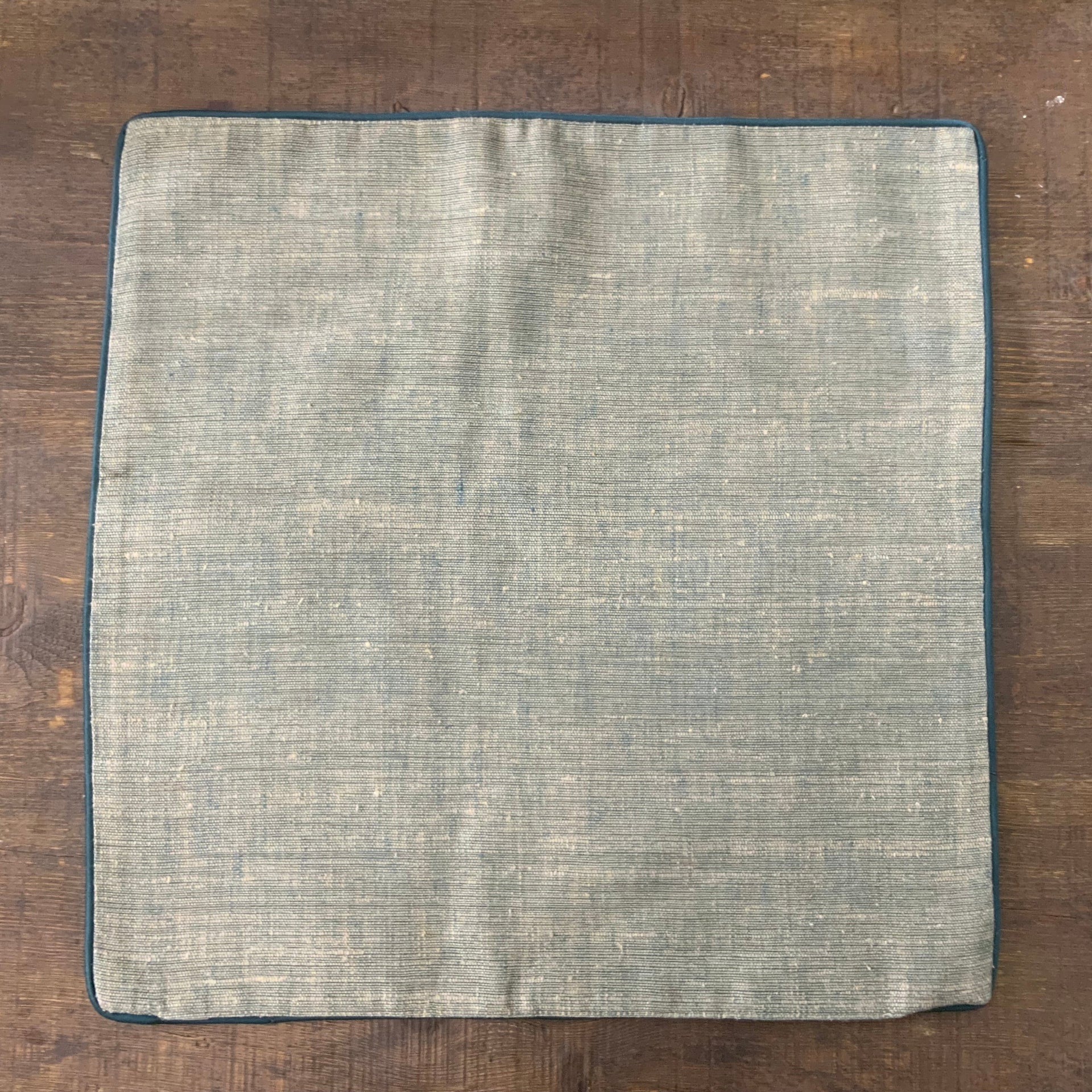 Square sage green cushion cover with a heavy slub texture and dark teal piped edges, organic woven fabric for minimalist home decor.