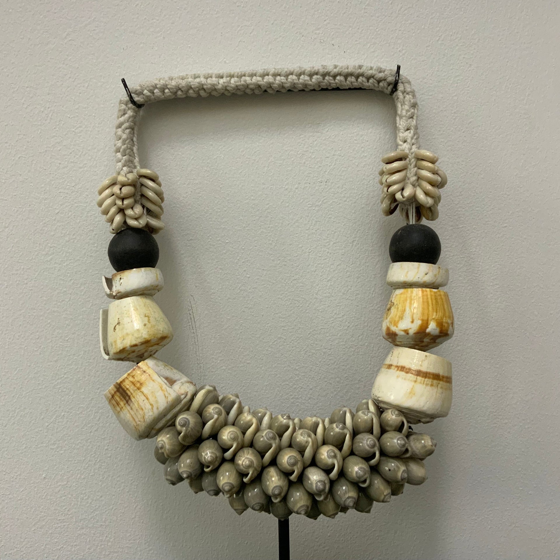 A handcrafted tribal-style statement necklace made of white sea shells, polished bone-colored resin rings, and black beads, displayed on a minimalist black metal stand