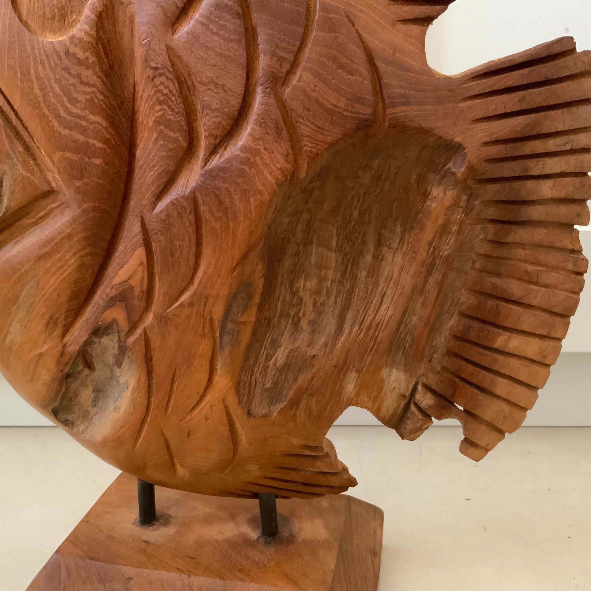Close-up of a carved wooden fish sculpture showing the natural wood grain, chiseled textures, and the dual metal rod mounting on a solid wood base.